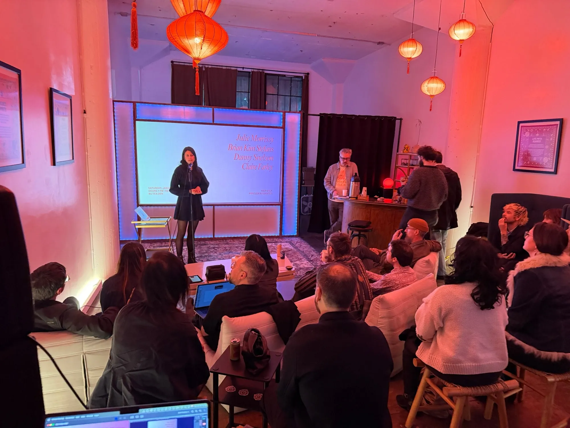 A poet reading at the mic in front of the projection wall, guests on lounge seats